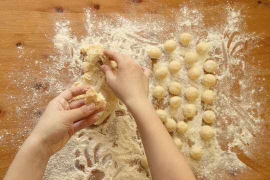 Children's hands tear off a piece of dough from a large bush against the background of many small balls for dumplings against the background of a wooden table showered with flour
