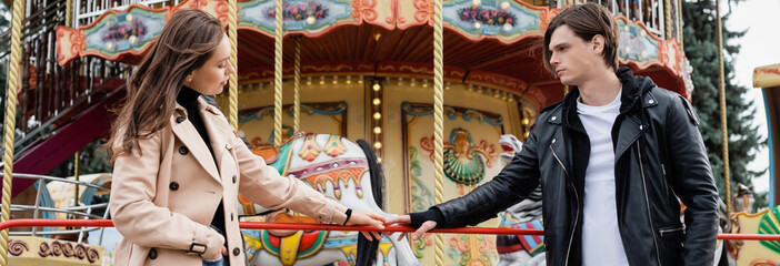 young couple standing near carousel and touching hands in amusement park, banner.