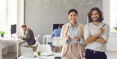 Happy smiling young business people in modern office workplace. Banner background with portrait of two satisfied successful colleagues, team leaders, teammates, business partners and friends at work - obrazy, fototapety, plakaty