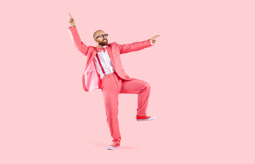 Funny dancer in pink party suit having fun in modern studio. Happy cheerful bald man with ginger beard, wearing funky suit, bowtie, trainer shoes and glasses dancing isolated on pink colour background