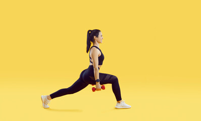Happy athlete enjoying fitness workout. Profile view of sporty fit attractive young woman in black leggings and crop top holding dumbbells, smiling and doing forward lunges exercise. Sport concept