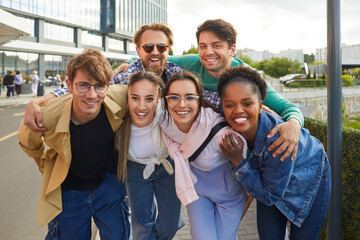 Portrait of happy carefree multiracial young friends posing together against backdrop of city street. Happy stylish casual men and women walking together smiling looking at camera. Friendship concept.