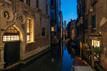 Venetian canal at night time, with the lights of the buildings reflecting in the calm waters