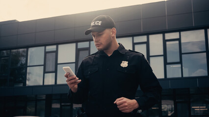 policeman in uniform and badge using smartphone on urban street.