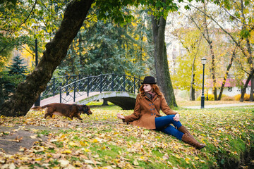 Young red-haired woman plays in the park near a small bridge with a red dachshund dog. 