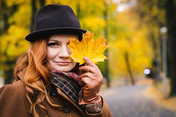 Portrait of a young red-haired woman in a hat against the background of autumn foliage. Woman posing covering one eye with a maple leaf