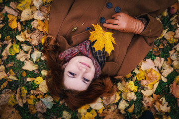Portrait of a young red-haired woman against the background of autumn foliage. Woman posing while lying on the grass and holding a leaflet in her hand, top view