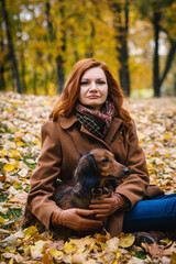 Young red-haired woman with a red dachshund dog sit on a ground in autumn in the park and looks into camera. 