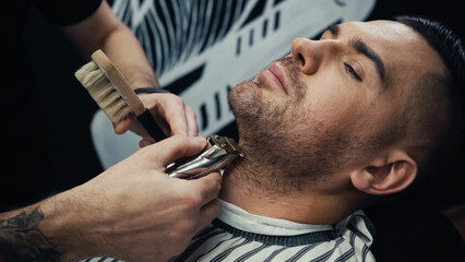 Barber trimming beard of customer in blurred barbershop.