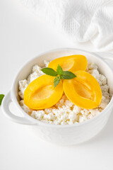 Fresh cottage cheese in white bowl with mint leaves and peach pieces on white background. Copy space