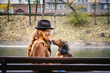 Young red-haired woman sit on a bench in autumn in the park with a red dachshund dog. Profile view