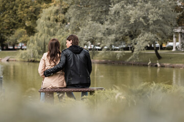 back view of young man in autumnal outfit hugging girlfriend while sitting on bench near lake in park.