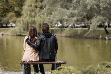 back view of young man in black jacket hugging girlfriend while sitting on bench near lake in park.
