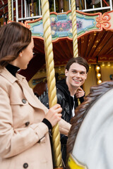 happy young man looking at girlfriend riding carousel horse in amusement park.