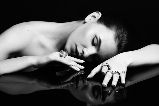 Fashion and make-up concept. Studio portrait of beautiful woman with fancy make-up and many various rings on her fingers lying on black glass surface. Model with closed eyes. Black and white image