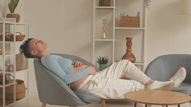Short haired young pregnant woman with hands on her belly relaxing in armchair at home