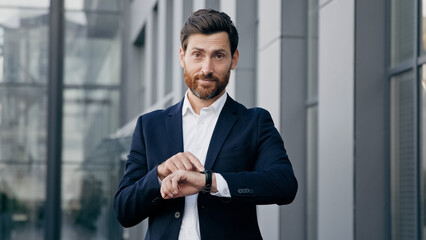 Professional confident businessman standing outdoors waiting for meeting looks at wristwatch checking time displeased caucasian man pointing on watch looking at camera dissatisfied with being late