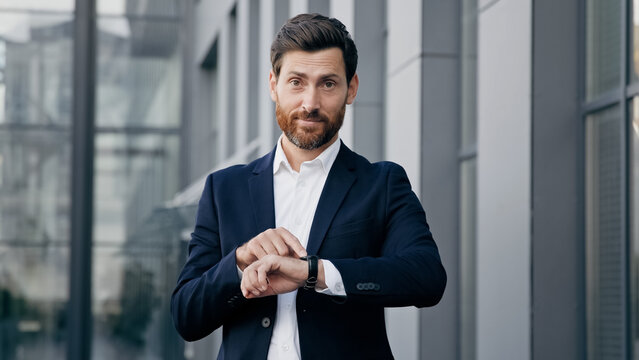 Professional confident businessman standing outdoors waiting for meeting looks at wristwatch checking time displeased caucasian man pointing on watch looking at camera dissatisfied with being late - Powered by Adobe