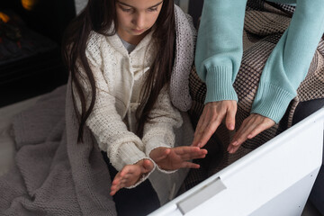 Child warming hands near electric heater at home, closeup