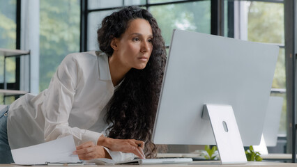 Young attractive hispanic woman professional manager developer working in modern office focused businesswoman checking electronic information on computer using paper documents researching sales data