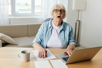 a shocked elderly lady is sitting at home in a cozy room at her desk with a laptop and looks up with her mouth wide open, thinking about solving work problems. The concept of working from home
