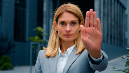 Serious woman standing outdoors looking at camera showing stop gesture holding palm in front...