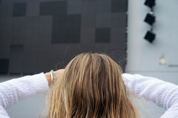 Fototapeta premium Hinterkopf von junger Frau mit blonden langen Haaren, weißem Pullover und verschränkten Armen vor Kopf steht vor großer weißer Fassade mit schwarzer Leinwand auf Platz in Stadt am Morgen im Sommer