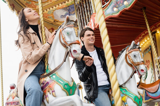 happy young couple holding hands and riding carousel horses in amusement park.