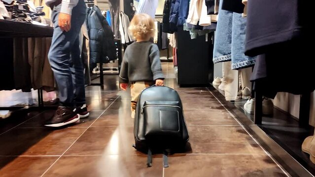 A cute little two-year-old child with a backpack goes shopping on his own surrounded by a lot of strangers and a forest of their feet. Funny footage, a concept for advertising a clothing store