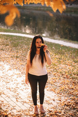 Young thoughtful stylish woman holding sunglasses and yellow leaf while standing in park during sunny weather in autumn
