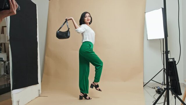 Full shot of Asian model in elegant outfit holding black leather bag, jumping and posing on camera in photo studio while shooting clothing for fashion lookbook