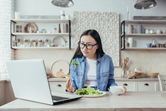 An interested Asian woman is sitting at the kitchen table at home. Carefully watches the online seminar on dietetics and healthy eating. He eats a fresh salad, holds a cup with a drink in his hand.