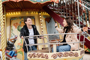 Fototapeta na wymiar cheerful man in stylish outfit looking at happy girlfriend laughing on carousel in amusement park.