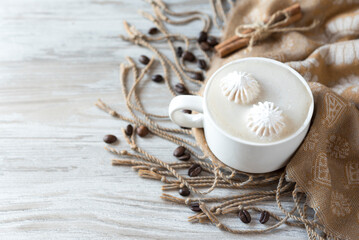 Cup of coffee with two small merengues on a top, some scattered coffee beans and beige fabric scarf on the light wooden background. Flat lay. Top view. Cozy composition