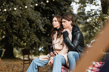Fototapeta premium stylish man in jacket hugging young girlfriend in beige coat sitting in park.