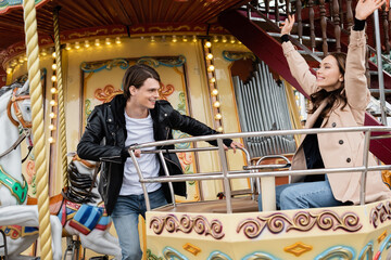 cheerful man in trendy outfit looking at happy girlfriend with raised hands on carousel in amusement park.