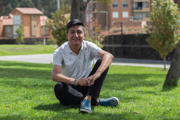 Cheerful young Latin man sitting on the green grass of a park on a sunny day while looking at the camera