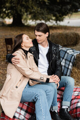 Fototapeta premium stylish man in black jacket hugging girlfriend in trench coat while sitting on blanket in park.