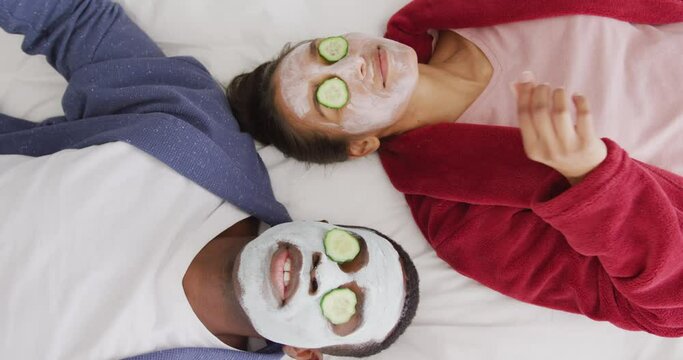 Video of happy diverse couple wearing cleansing face masks and cucumber on eyes lying on bed smiling