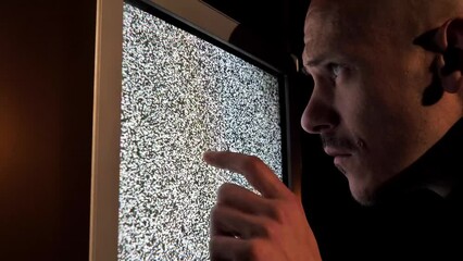 A man with a puzzled expression is trying to fix a broken old TV. Close-up. Surreal zombie TV scene, media manipulation and propaganda. Glitch interference and static noise on the screen. TV drugs