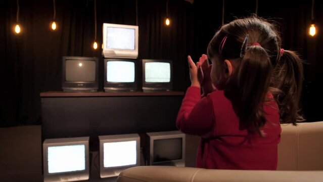 A little girl sits in front of a lot of TVs with glitch interference and claps her hands. The child watches non-working TVs. Zombie TV concept. Propaganda and manipulation from television. Zombie box.
