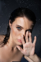 Young brunette woman with makeup touching wet glass on grey background.