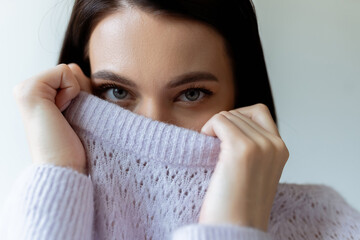 young brunette woman looking at camera while obscuring face with soft knitted sweater isolated on...