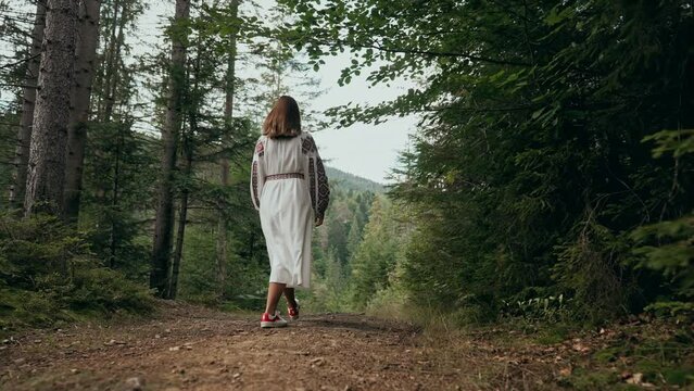 Back view of unrecognizable ukrainian woman walking in fir forest, Carpathian mountains nature. Girl in traditional embroidery vyshyvanka dress. Ukraine, freedom, ethnic national costume