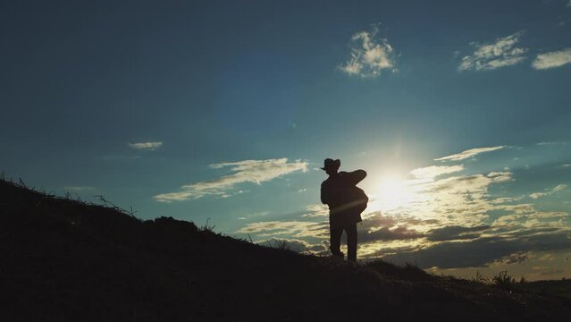 Sportive man walks in highland at bright back sunset light exploring wild nature in evening. Dark silhouette of male person in hat walking in rural area against illuminated cloudy sky backside view