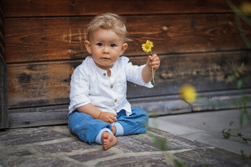 one year old happy blond baby boy in white shirt sitting outside on the ground infront of a rustic...