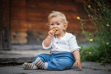 Fototapeta premium one year old happy blond baby boy in white shirt sitting outside on the ground infront of a rustic wooden hut and holding a yellow marguerite flower to send love and happiness