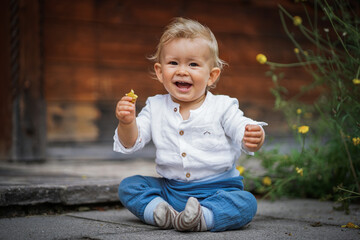 one year old happy blond baby boy in white shirt sitting outside on the ground infront of a rustic...