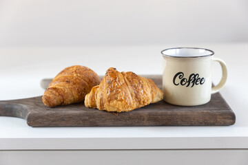 Two croissants, a cup with coffee inscription on a wooden board on a white table. Place for text