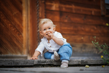 one year old happy blond baby boy in white shirt sitting outside on the ground infront of a rustic wooden hut and holding a yellow marguerite flower to send love and happiness - obrazy, fototapety, plakaty
